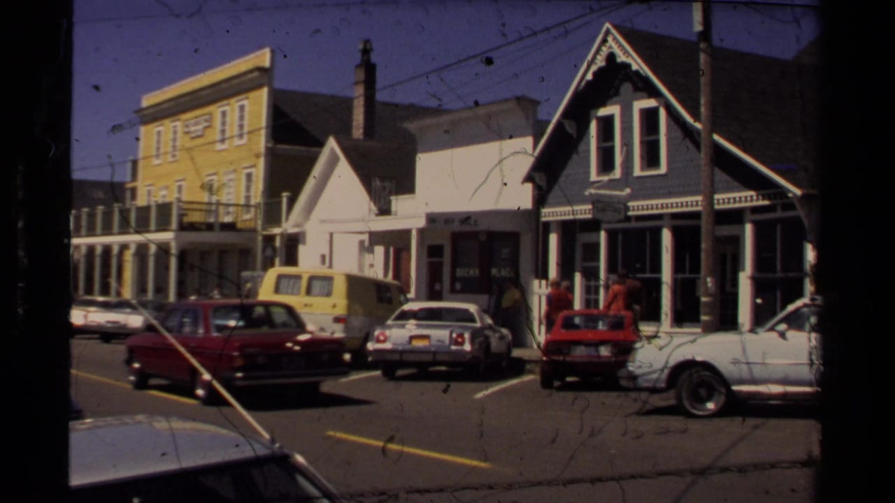 Mendocino downtown street view with Victorian-style storefronts, 1980s parked cars, a yellow van, and pedestrians near shopfronts.