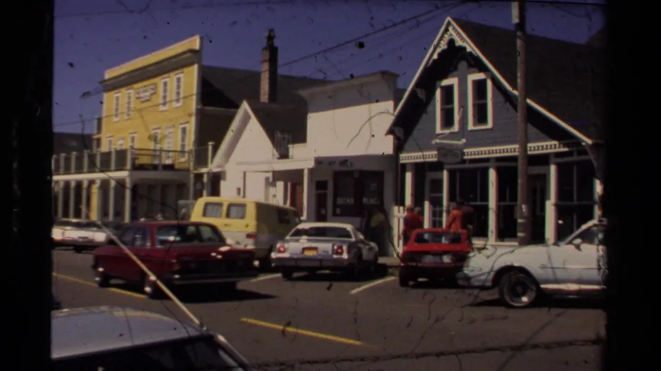 Mendocino downtown street view with Victorian-style storefronts, 1980s parked cars, a yellow van, and pedestrians near shopfronts.