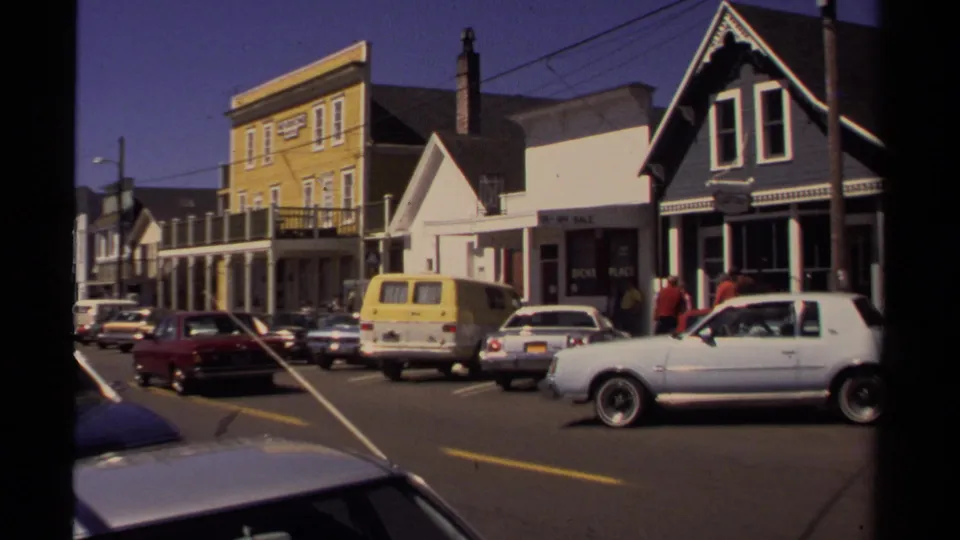 Mendocino street scene with colorful commercial buildings, angled parking, a yellow van, pedestrians, and a white car in the foreground.