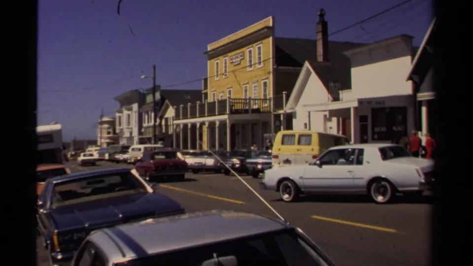 Wide Mendocino downtown view showing 1980s cars, a yellow van, storefronts, overhead wires, and pedestrians near the sidewalk.
