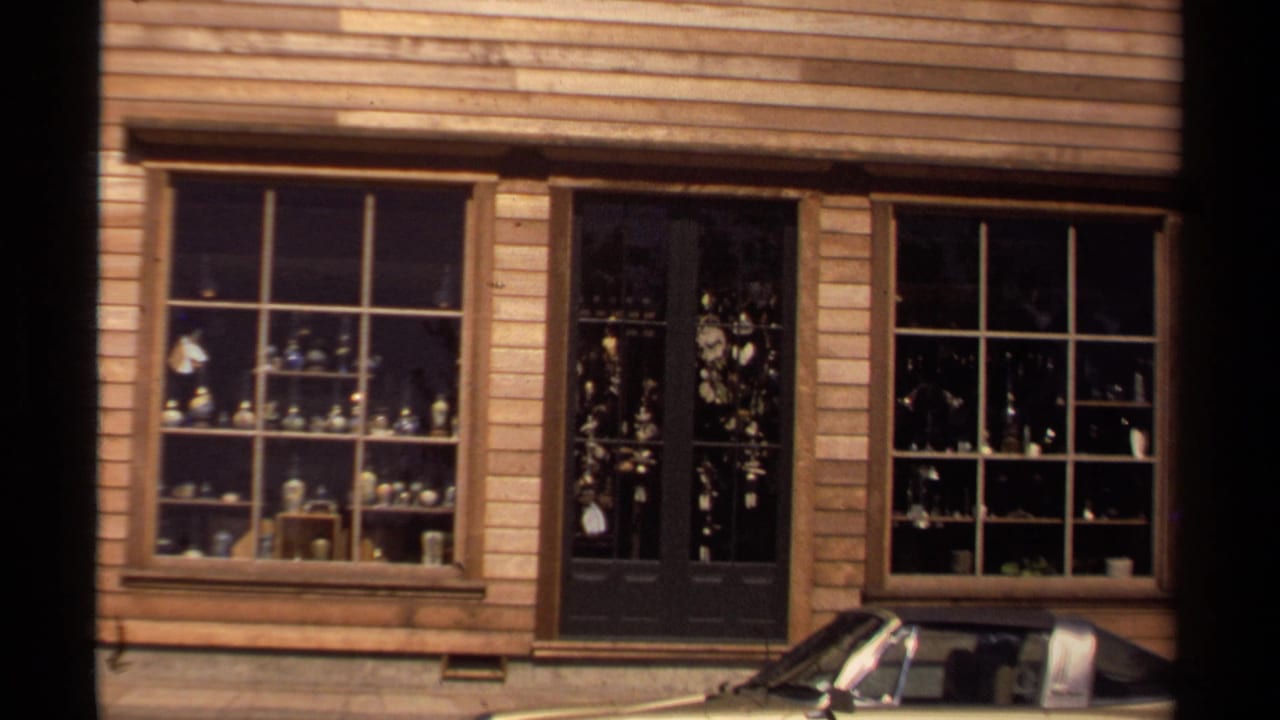 Shopfront windows and doorway with glassware displays and a parked car at the curb.