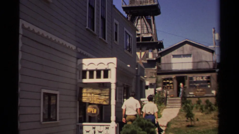 Mendocino small-town street scene with two pedestrians approaching shopfronts and mixed historic-looking facades.