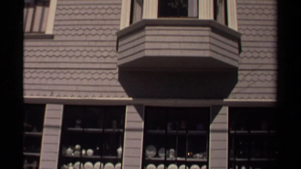 The camera frames the bay window and lower shop display in Mendocino, with shelves of white china visible through multi-pane windows.