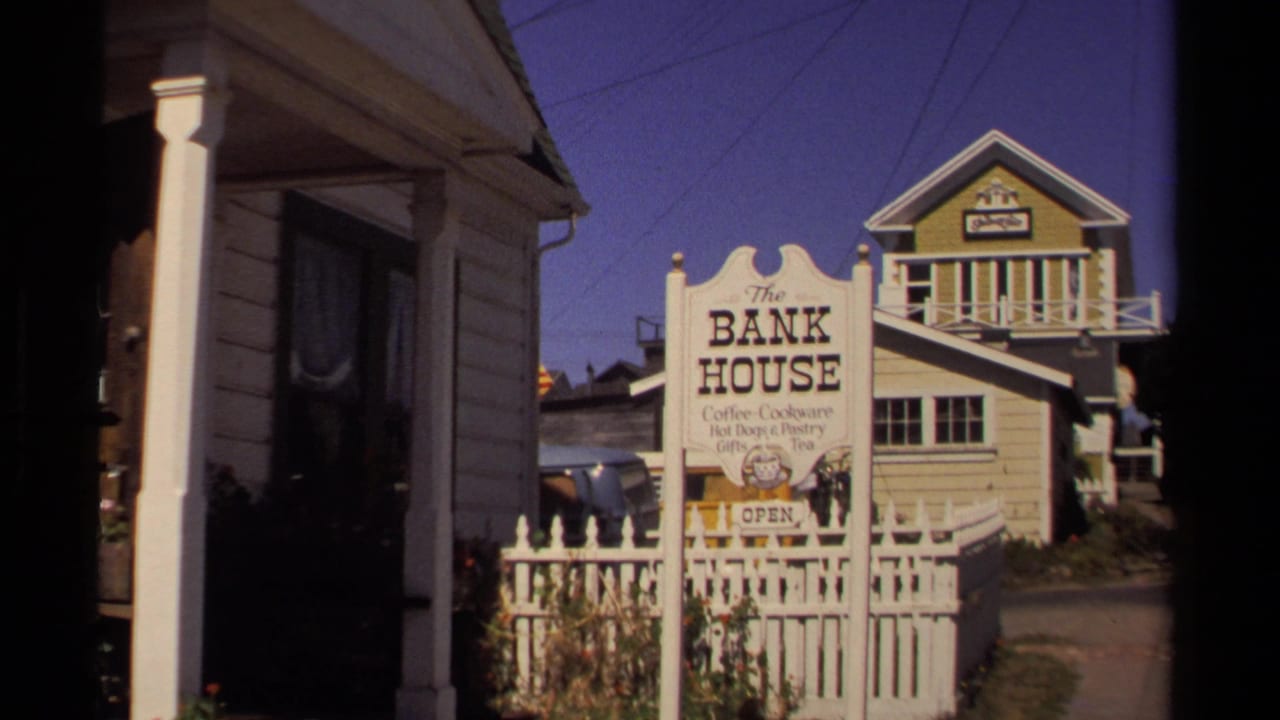 Mendocino, California 1980 frame showing The Bank House sign, a white fence, and wooden buildings along a narrow walkway.