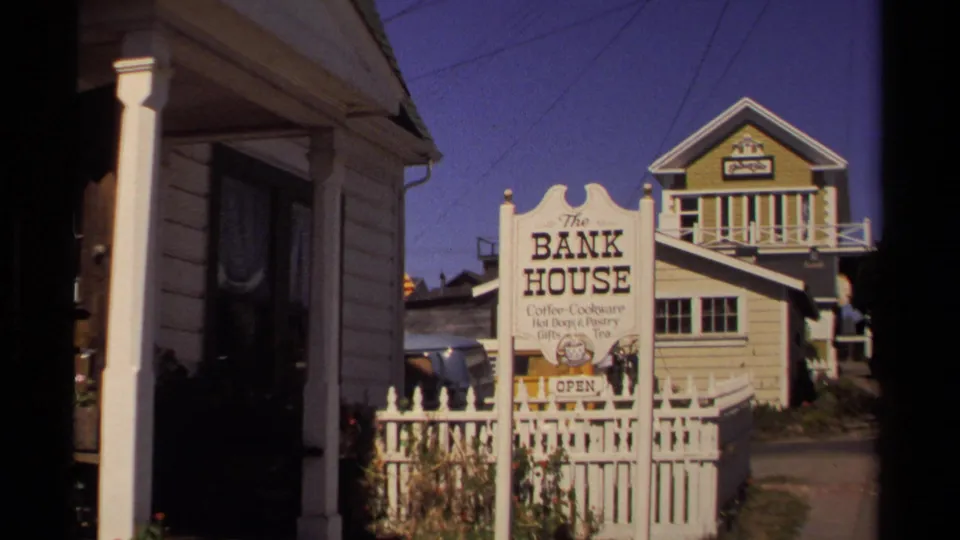 Mendocino, California 1980 frame showing The Bank House sign, a white fence, and wooden buildings along a narrow walkway.