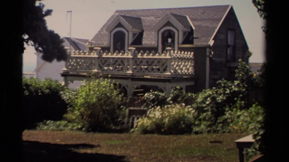Mendocino frame of a Victorian-style wooden house with decorative balcony railing, dormer windows, and dense garden plantings.