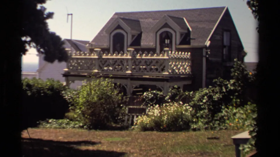 1980 Mendocino view of a wooden house with decorative balcony details, dormer windows, shrubs, flowers, and a small lawn.