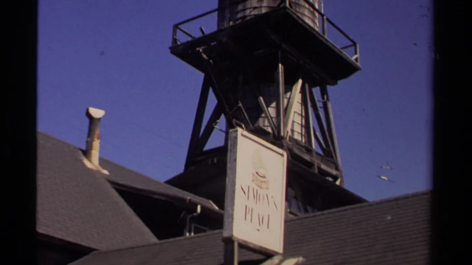 Mendocino, California 1980 frame showing a water tower structure above rooftops and a visible Simon's Place sign against blue sky.