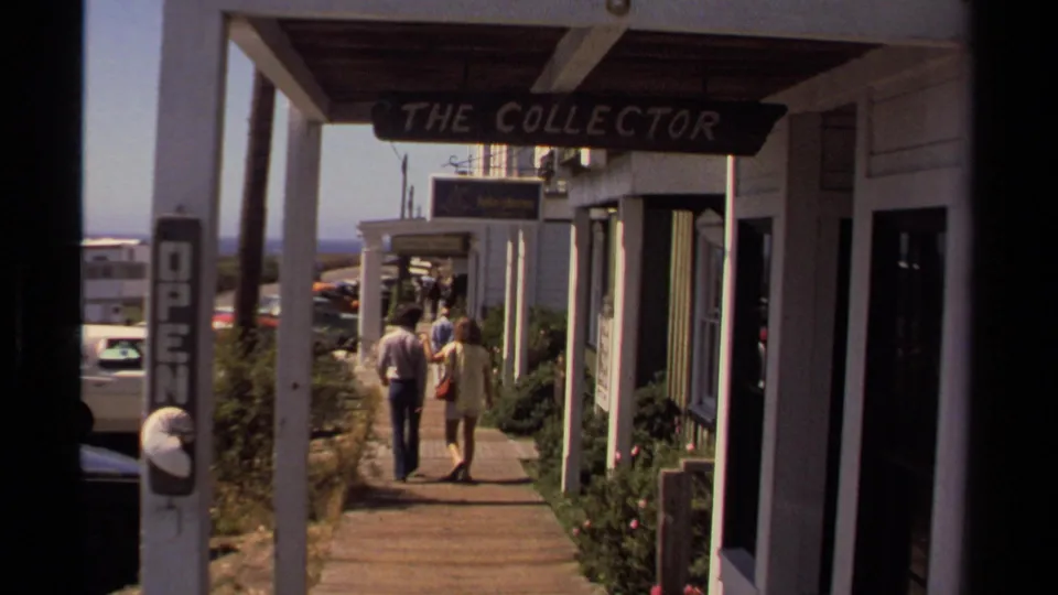 Two pedestrians walk along a covered Mendocino sidewalk past storefronts including a sign reading The Collector.