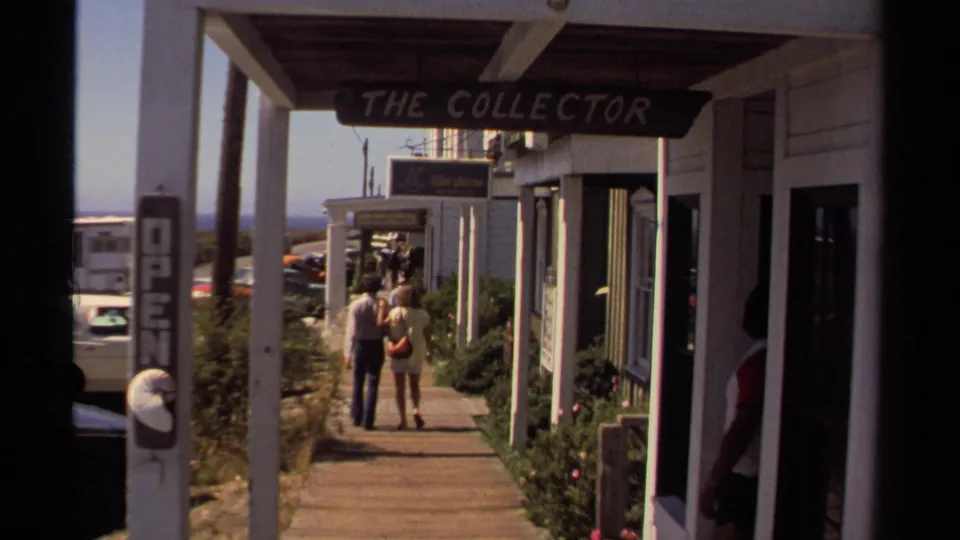 1980 Mendocino home-movie frame showing a couple walking away along a sunlit wooden boardwalk past local shops and signs.