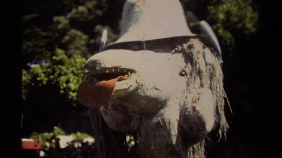 Mendocino 1980 home-movie frame showing the same grotesque animal mask on a pole against dark trees and garden foliage.