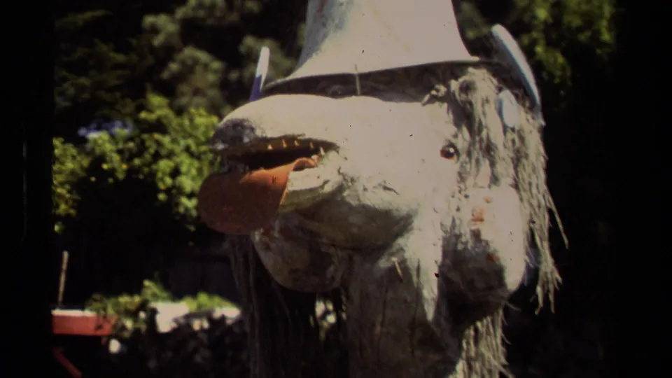 1980 Mendocino 8mm frame with a close view of a shaggy goat-like mask decoration, white hat, and sunlit plants behind it.