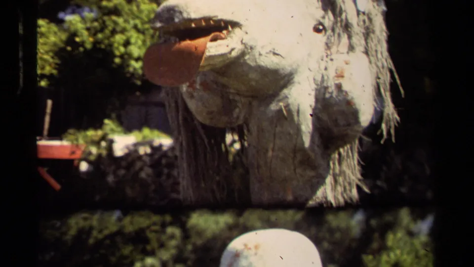 Final 1980 Mendocino frame showing the animal-like pole-mounted mask in bright sun, with green foliage and deep shade behind.
