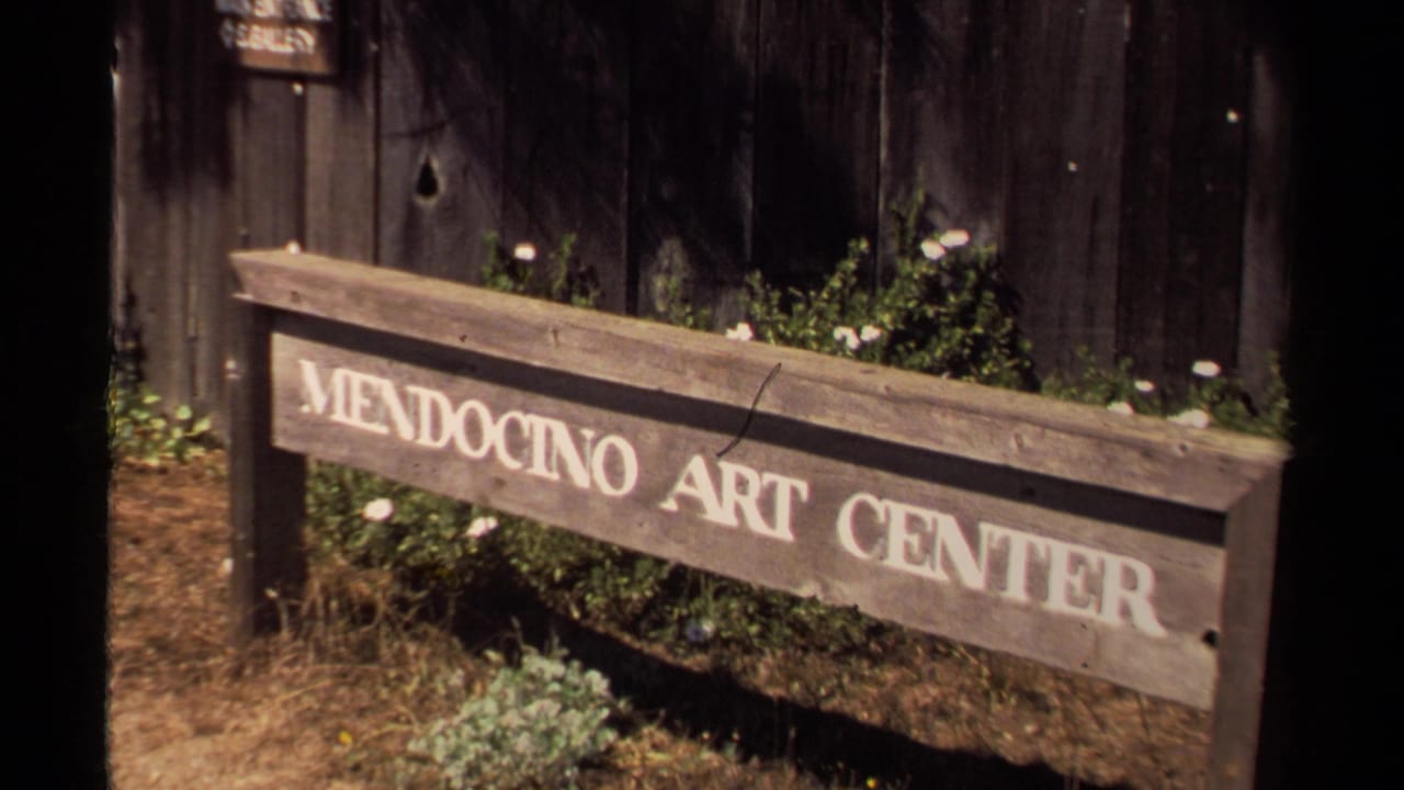Wooden sign reading Mendocino Art Center beside a dark building and plants.