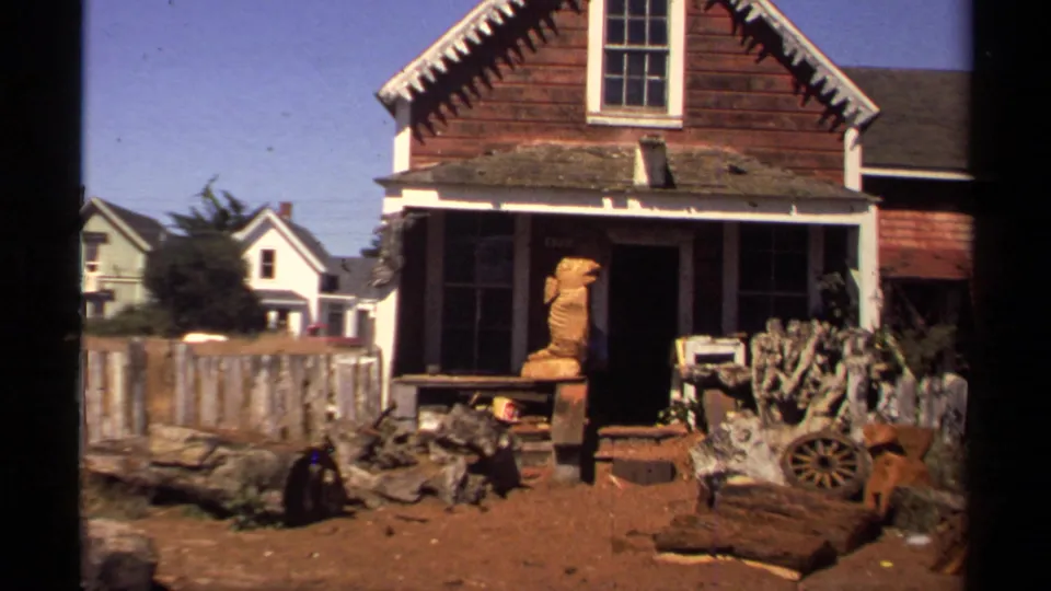 1980 Mendocino frame of a weathered wooden building with a porch, fence, yard objects, cactus-like plants, and a carved wooden figure outside.