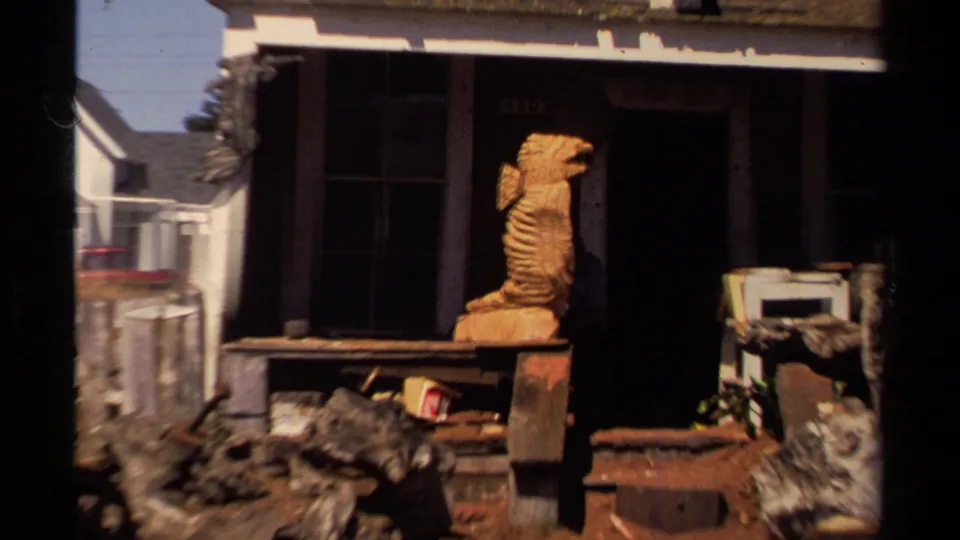 Close 1980 Mendocino home-movie view of a carved wooden figure near a dark porch, with weathered objects and a fence in the foreground.
