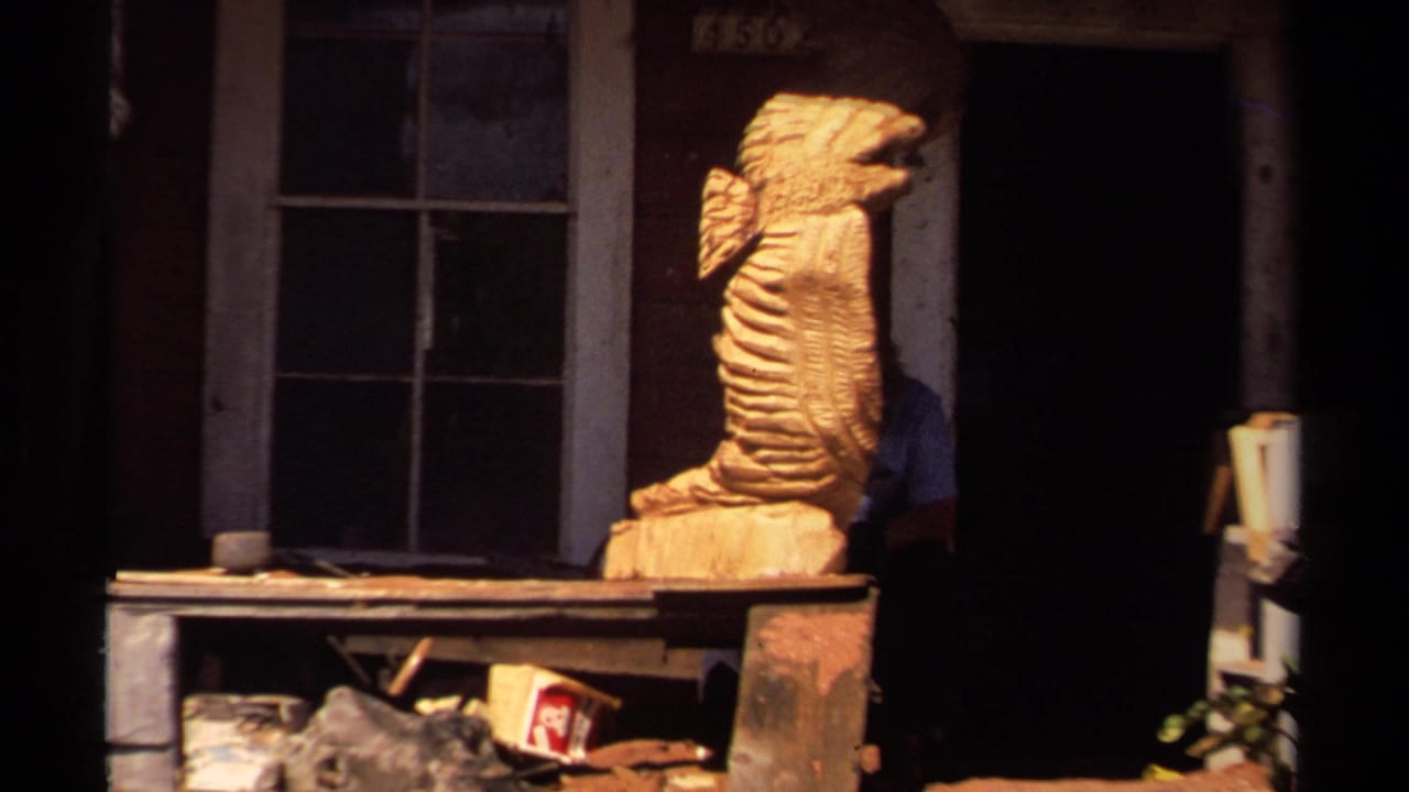 1980 Mendocino home-movie frame showing a large carved wooden sea creature on a porch, with windows, shelves, and deep doorway shadows.