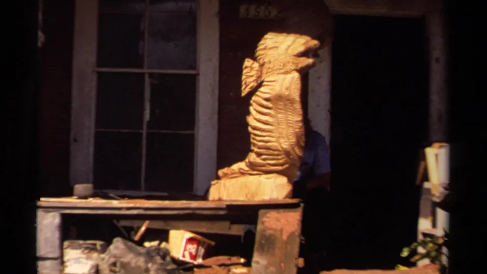 1980 Mendocino home-movie frame showing a large carved wooden sea creature on a porch, with windows, shelves, and deep doorway shadows.