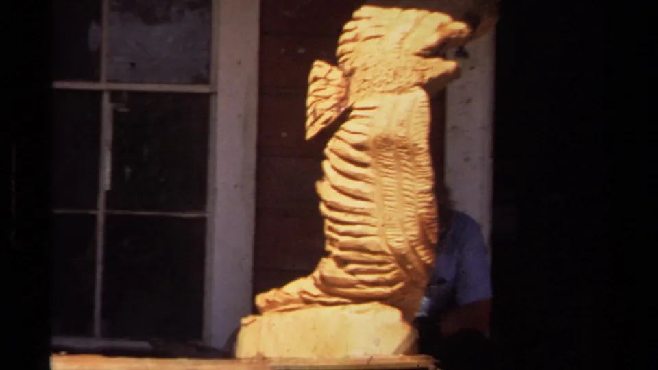 Closer 8mm frame of the Mendocino porch carving, with a partially visible person behind the wooden form and window trim in shadow.