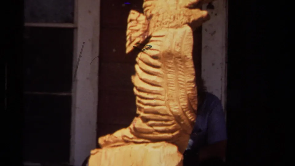 Tight 1980 home-movie view of the carved wooden form on a porch, cropped close against a window frame and dark entry area.