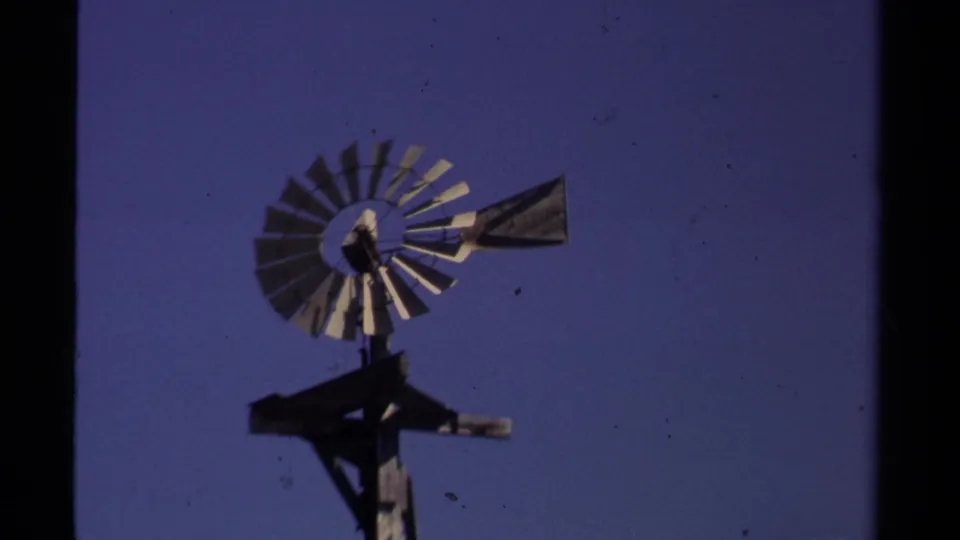 1980 Mendocino home-movie frame showing a metal windmill and wooden support silhouetted against a blue sky.