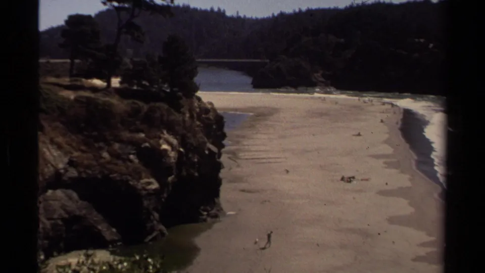 Mendocino coastal overlook showing a sandy beach, river mouth, rocky bluff, trees, surf edge, and small beachgoers in 1980 home-movie footage.