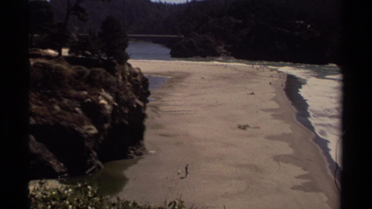 Elevated view of a sandy beach with a lone walker, rocks, surf, and distant hillside.