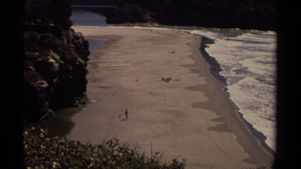 1980 Mendocino coastal frame with a lone beach walker, distant people, low surf, and rocky shoreline from an elevated view.