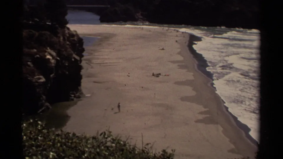 1980 Mendocino coastal frame with a lone beach walker, distant people, low surf, and rocky shoreline from an elevated view.