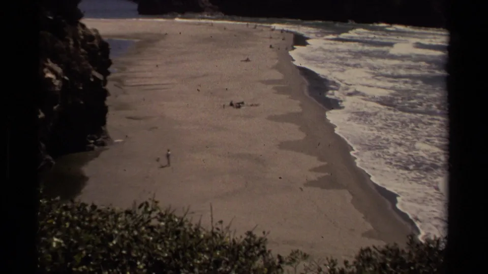 1980 Mendocino beach view showing a solitary walker near the lower sand, with surf, rocks, shrubs, and distant beach activity.