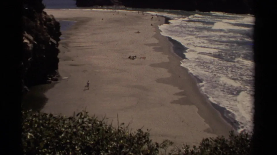 1980 Mendocino home-movie still of a lone person walking on a wide beach beside rolling Pacific surf and coastal rocks.