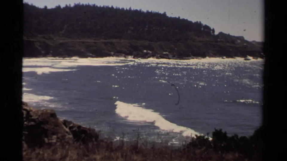 Sunlit Mendocino-area surf moves toward a rocky shoreline in grainy 1980 8mm footage.