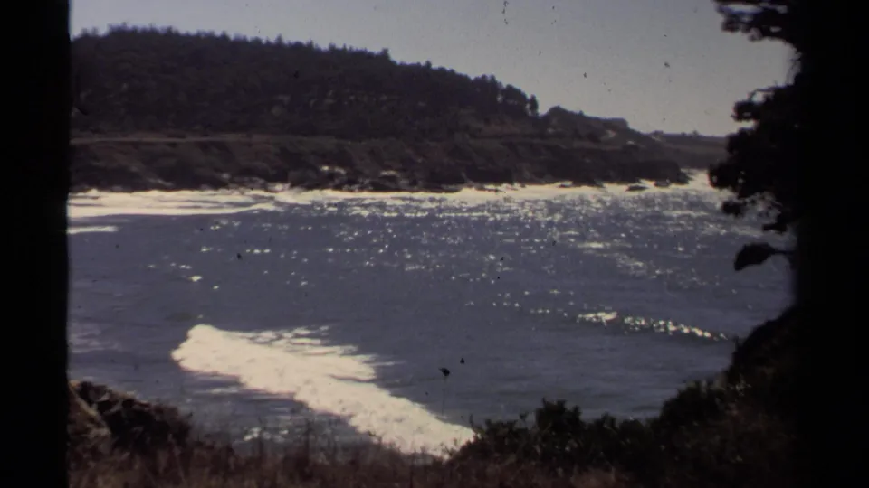A 1980 8mm view of the Mendocino-area coast shows sparkling water, white surf, rocks, and dark trees at frame edge.