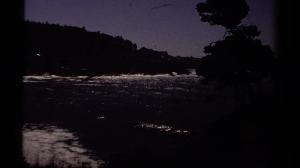 Underexposed 8mm view of the Mendocino shore, showing dark trees, bright surf bands, ocean water, and a wooded headland.