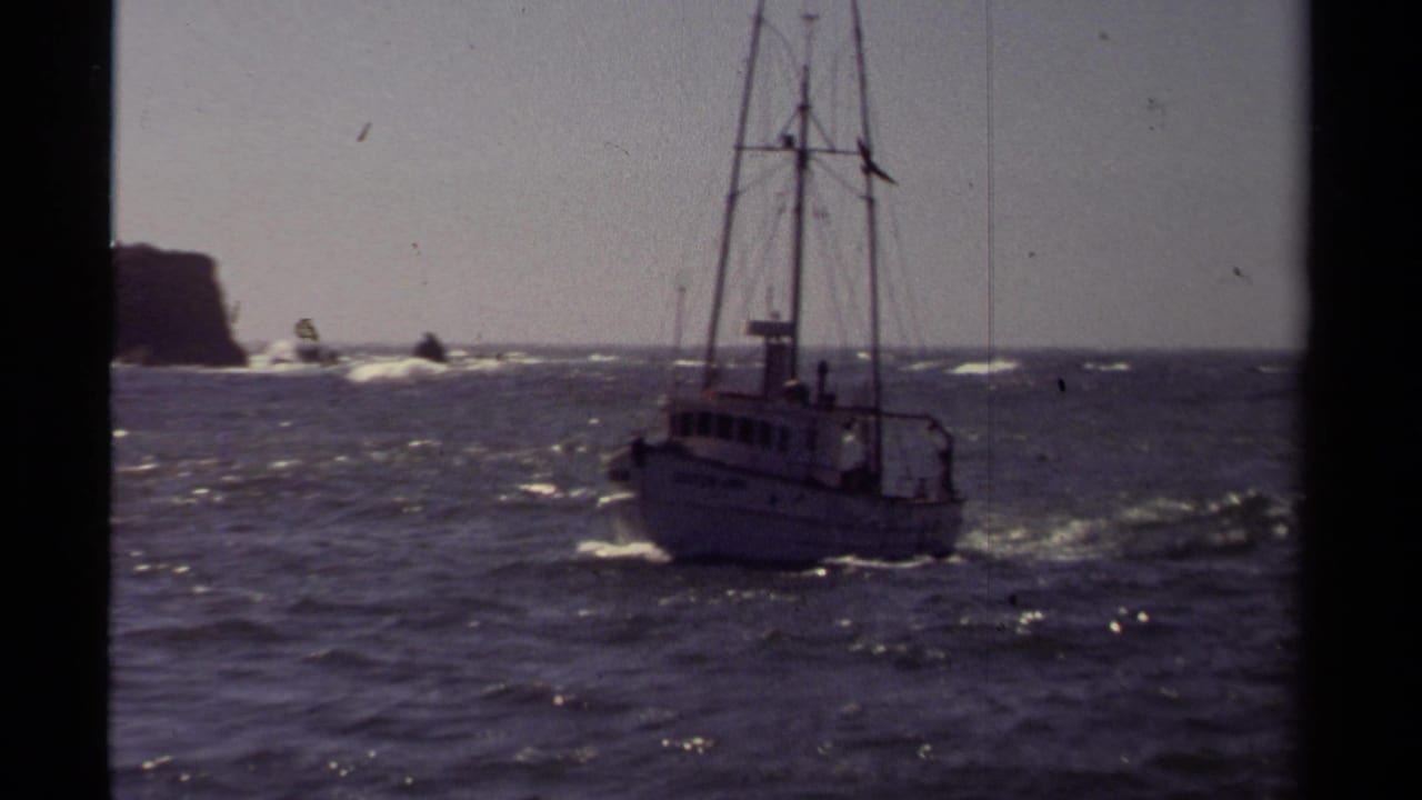 Fishing boat turns through choppy sea with rocks and surf in the distance.