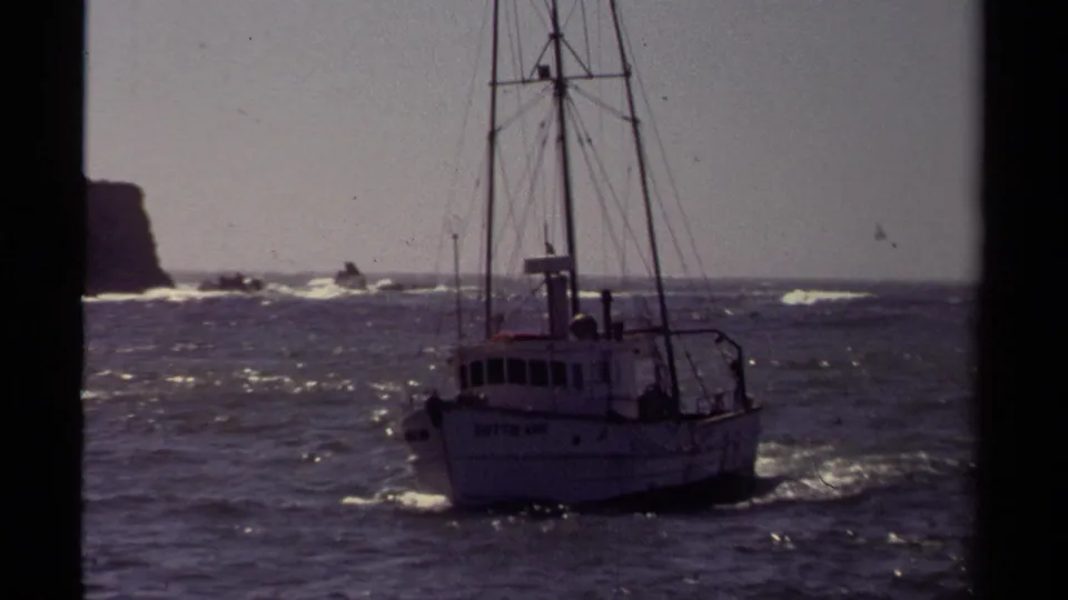 The white fishing boat continues past the camera in rough Mendocino water, with rigging, hull lettering, rocks, and surf visible.