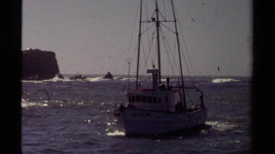 A small fishing boat moves through choppy Mendocino coastal water in 1980, with a rocky headland and surf behind it.