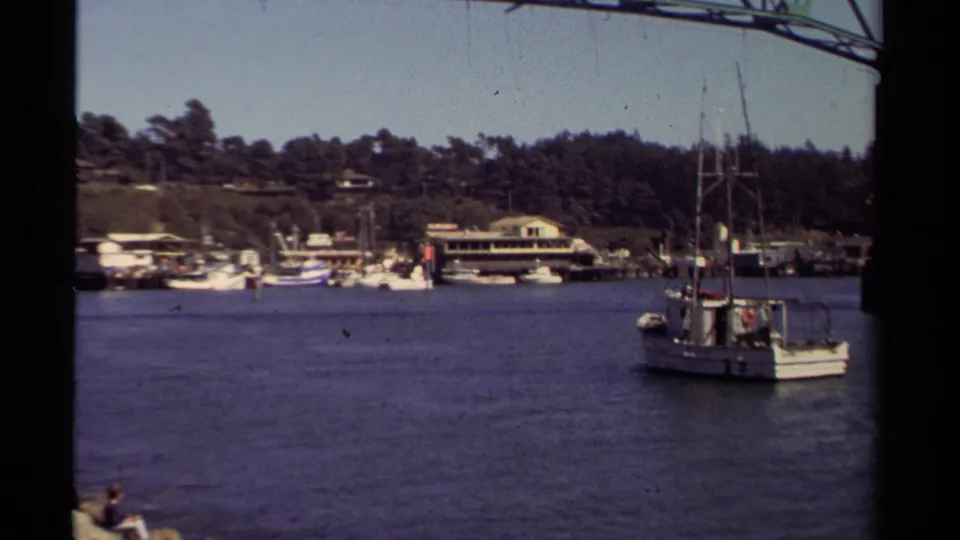 A wider Mendocino harbor view shows the fishing boat moving across calm water near docks, boats, buildings, and forested hills.