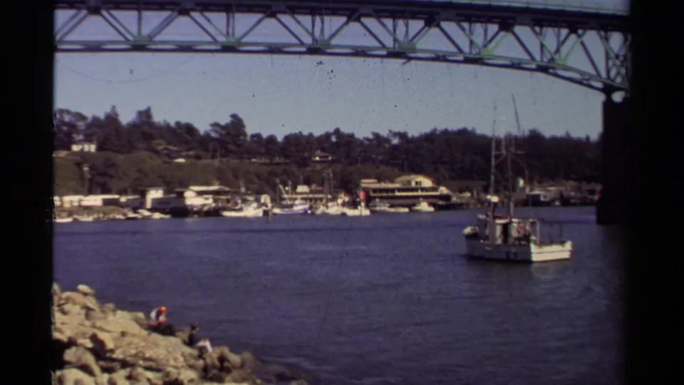 The boat passes near Mendocino harbor shore beneath a truss bridge in 1980, with rocks, distant people, and waterfront buildings visible.