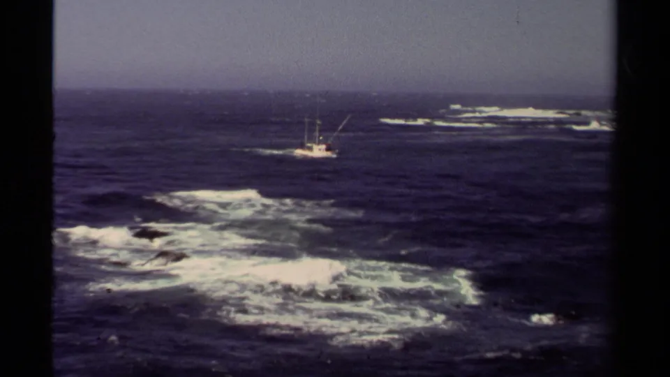1980 Mendocino-area 8mm frame of a fishing boat offshore, framed by foamy waves and a dark horizon.