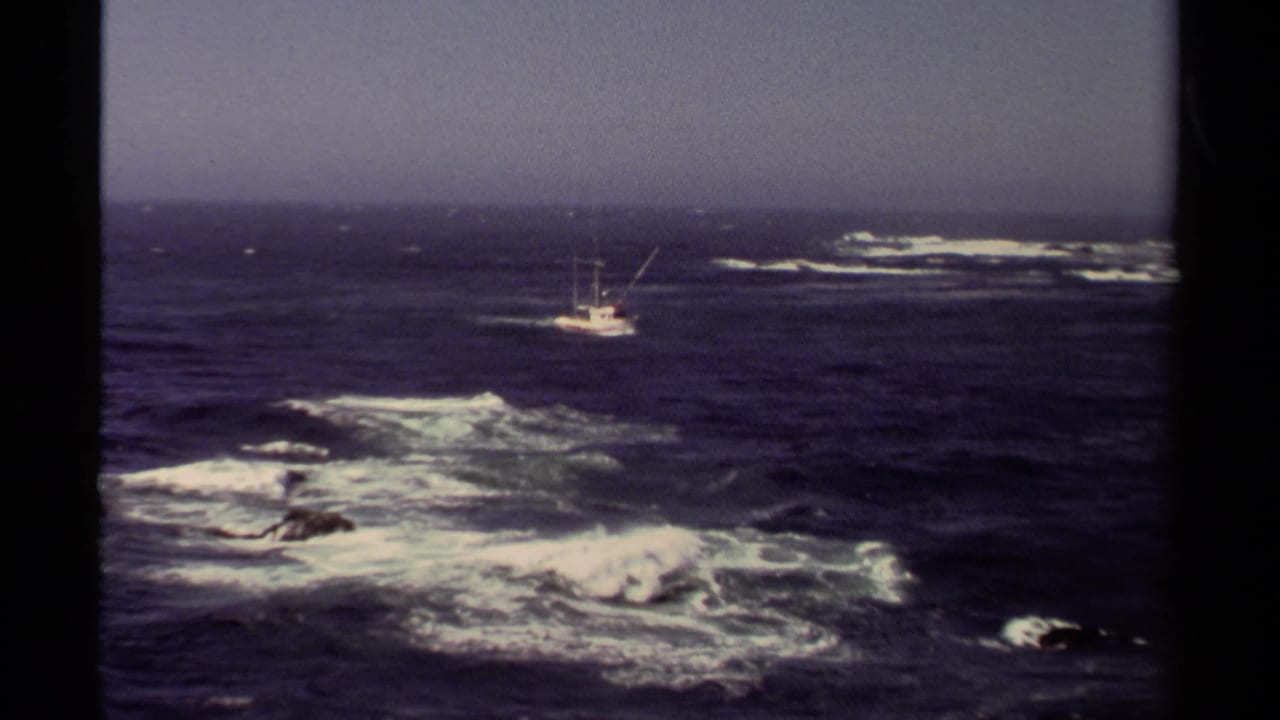 1980 8mm view of a white fishing boat off Mendocino, California, with dark blue water, whitecaps, and rocks in the foreground.