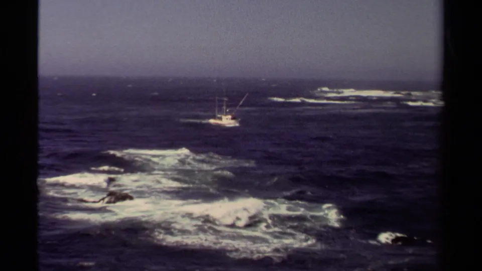 1980 8mm view of a white fishing boat off Mendocino, California, with dark blue water, whitecaps, and rocks in the foreground.