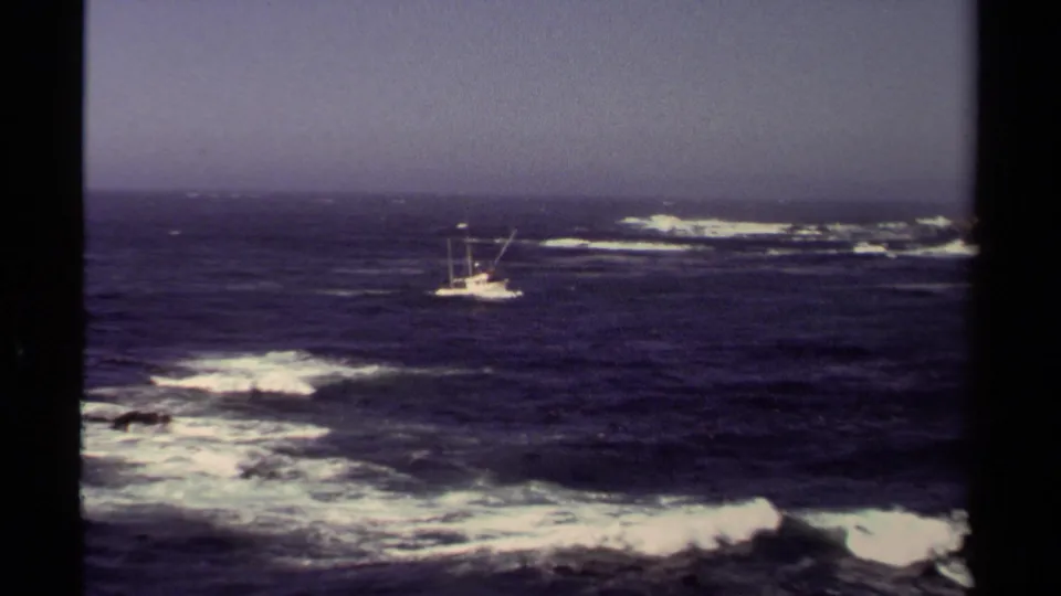 Mendocino coastal home-movie frame with a distant fishing boat, dark ocean, white surf, and a hazy sky.