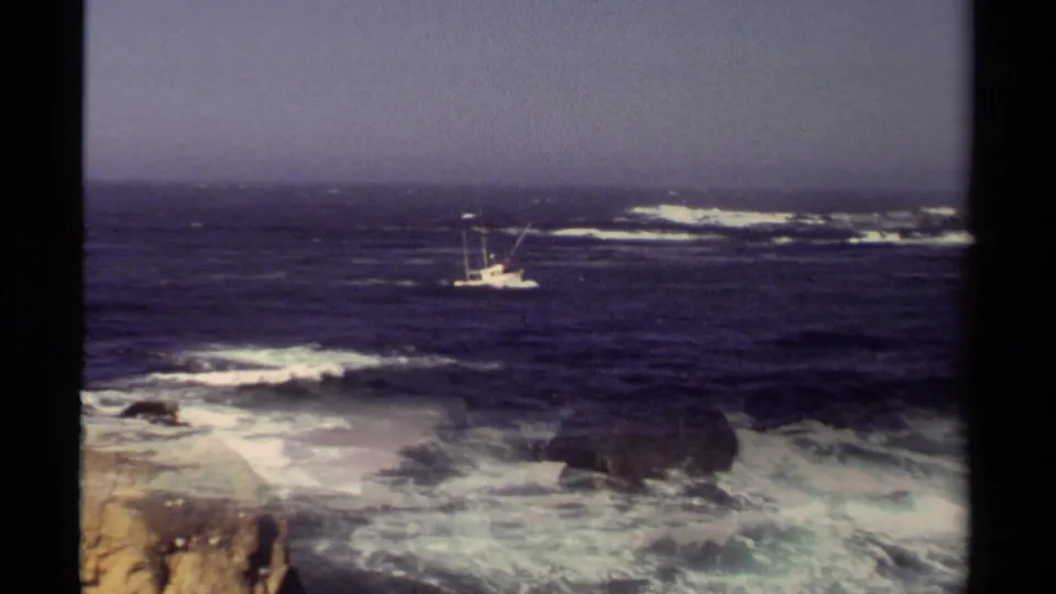 1980 Mendocino 8mm frame showing a small fishing boat beyond white surf breaking around coastal rocks.