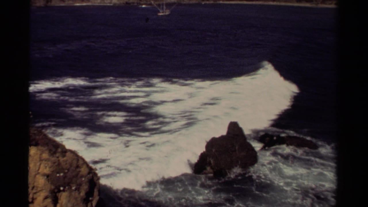 1980 Mendocino coastal frame with a broad white wave curling around dark rocks under muted light.