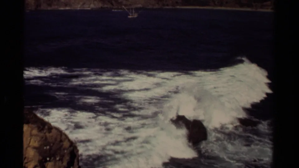1980 Mendocino 8mm frame showing rough Pacific surf surging over dark rocks near the shore.