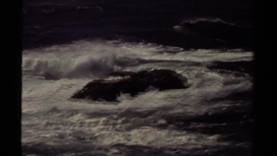 1980 Mendocino coastal home-movie frame showing rough surf breaking around dark rocks under dim overcast light.