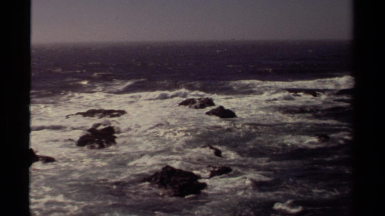 White water churns around scattered offshore rocks under a muted sky, bordered by dark vertical framing.