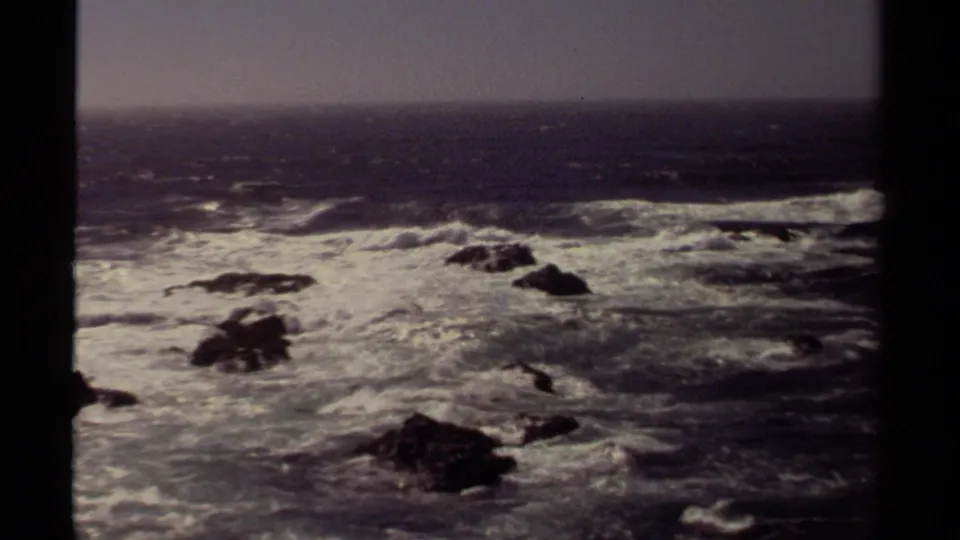 Vintage 8mm view of Mendocino coastline surf rolling across dark rocks under a gray, hazy sky.