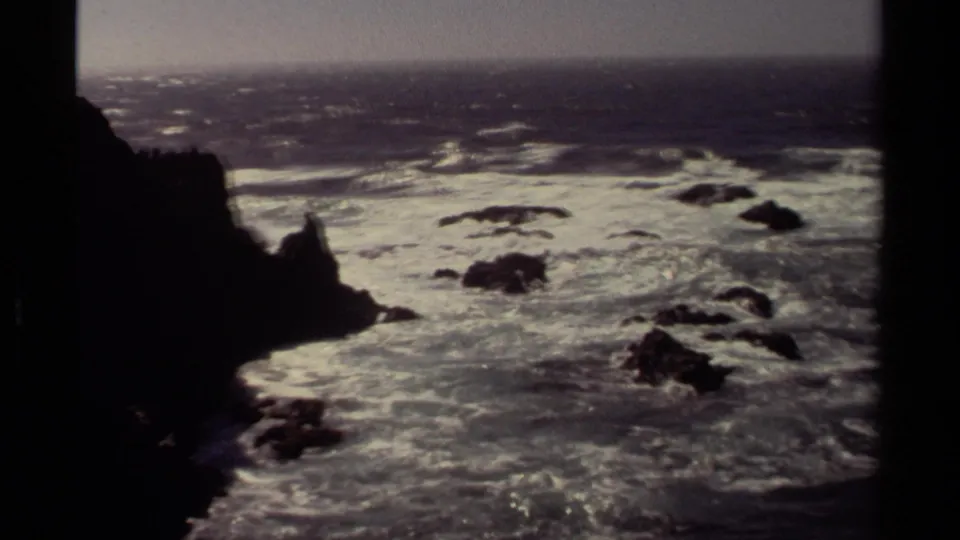 1980 Mendocino home-movie frame with a dark foreground rock silhouette and bright surf spread across the shoreline.